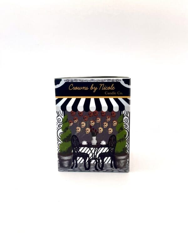 A Sass in Scarlett box from "crowns by nicole candle co." with a parisian café-themed design, featuring a black and white striped awning and a table with chairs.